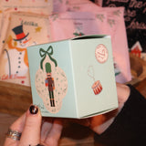 Someone holding the Nutcracker Bath Bomb in front of other holiday bath products, showing off the side.