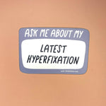 The Ask Me About My Latest Hyperfixation - Vinyl Sticker on a light orange background,