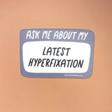 The Ask Me About My Latest Hyperfixation - Vinyl Sticker on a light orange background,