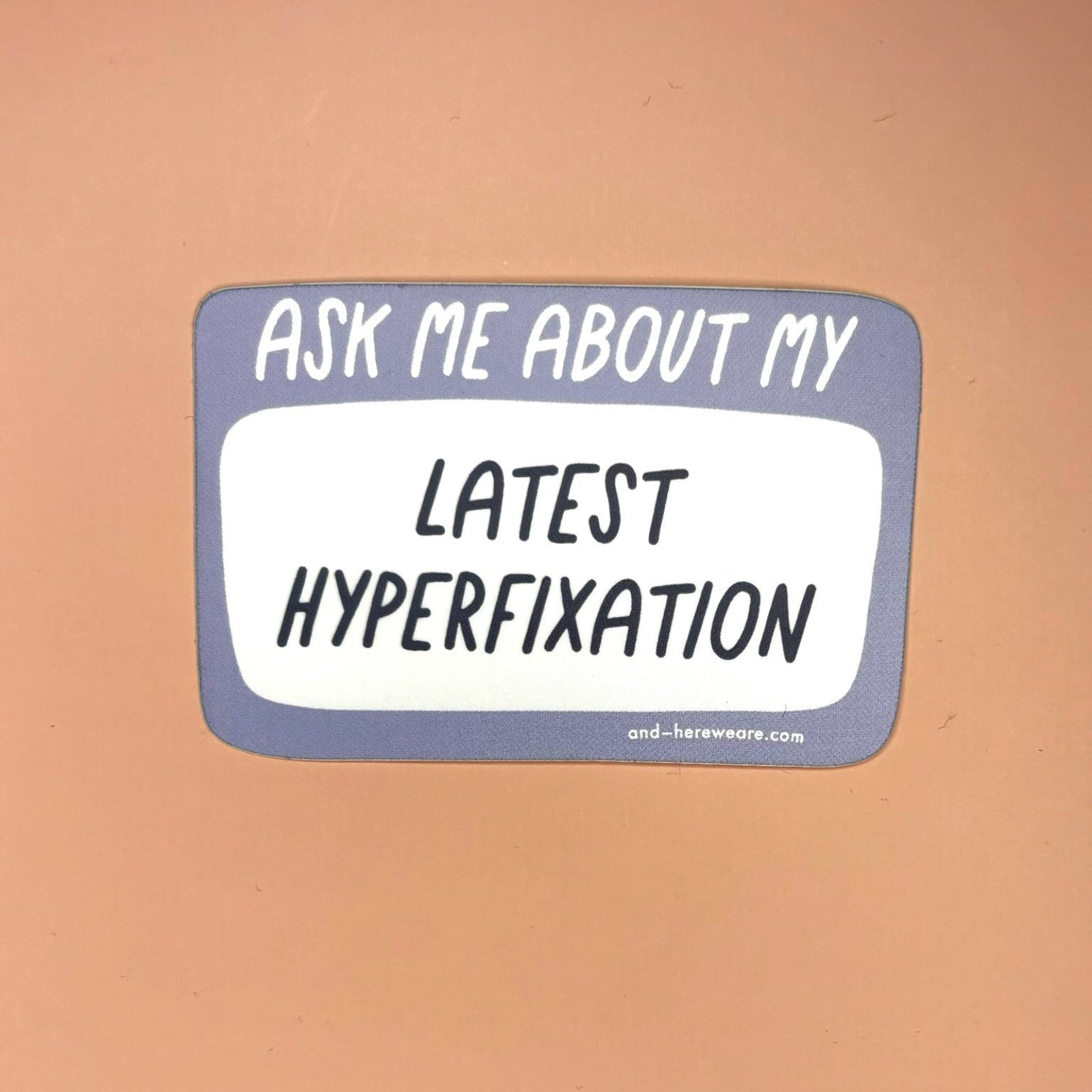 The Ask Me About My Latest Hyperfixation - Vinyl Sticker on a light orange background,