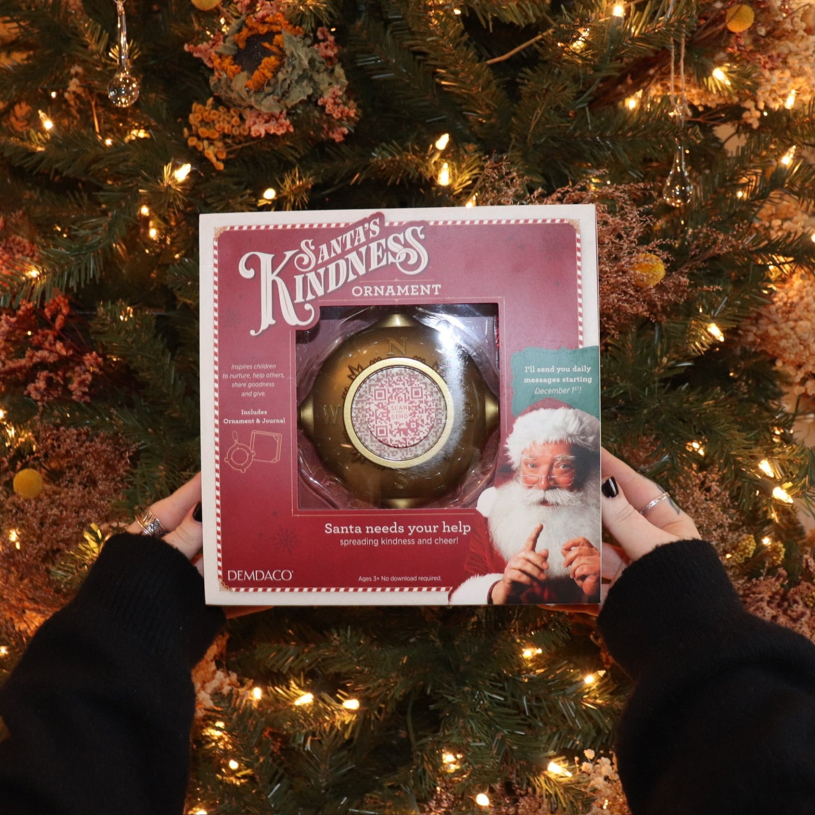 Someone holding the Santa's Kindness Ornament & Journal in front of a Christmas tree.