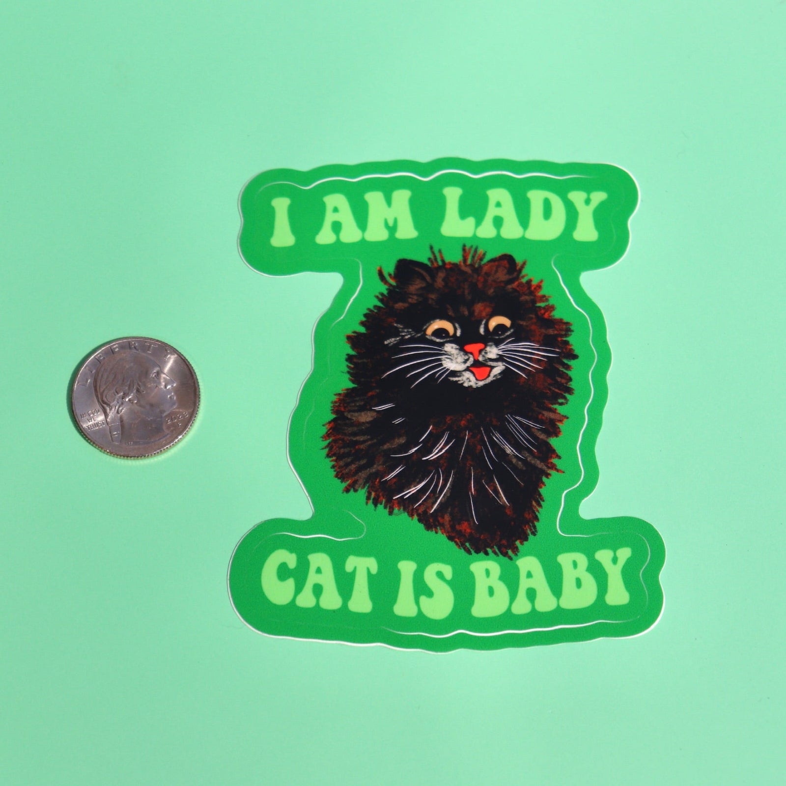 The I Am Lady Cat Is Baby 3 Inch Sticker next to a quarter on a green background.