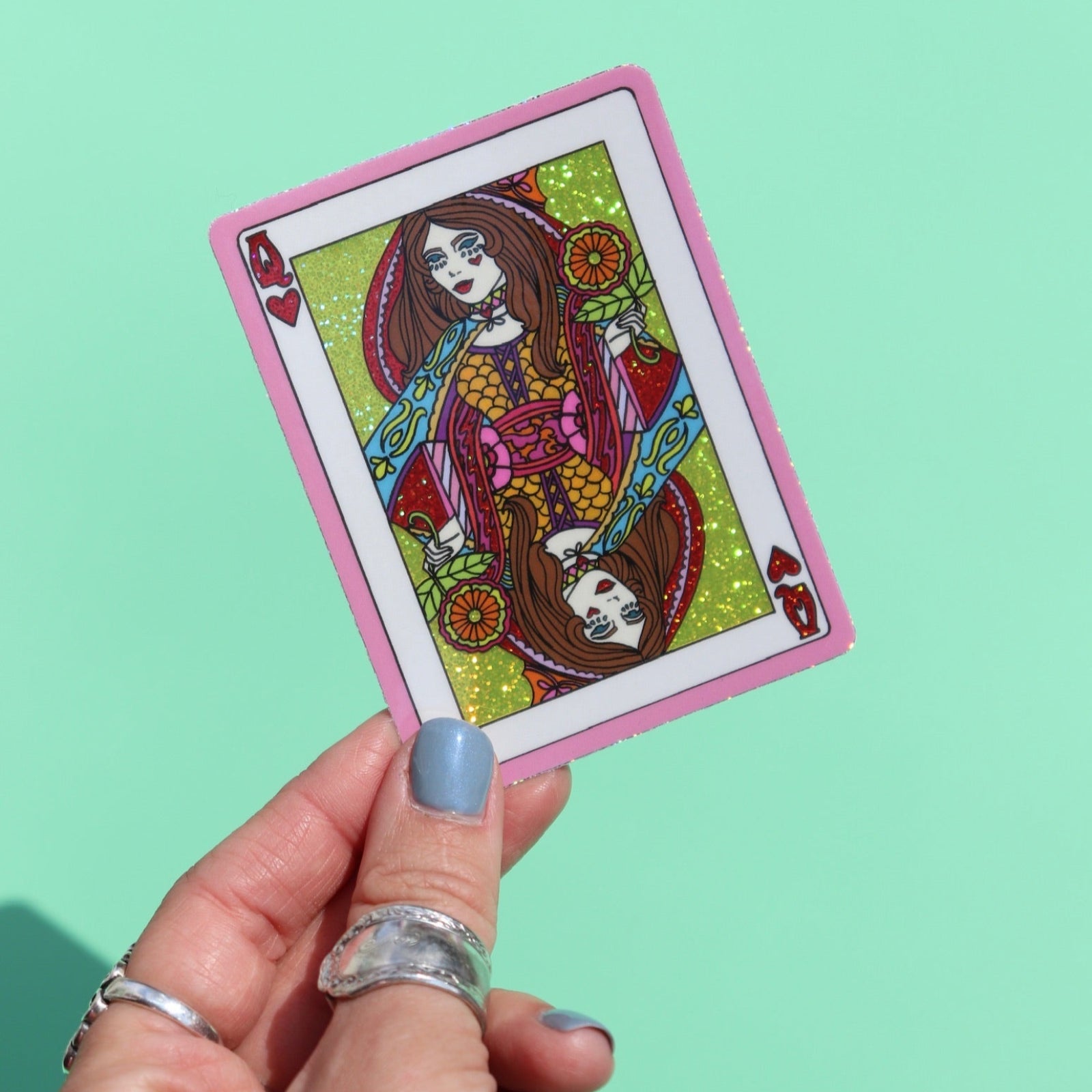 Someone holding the Queen of Hearts Glitter 3 Inch Sticker on a green background.