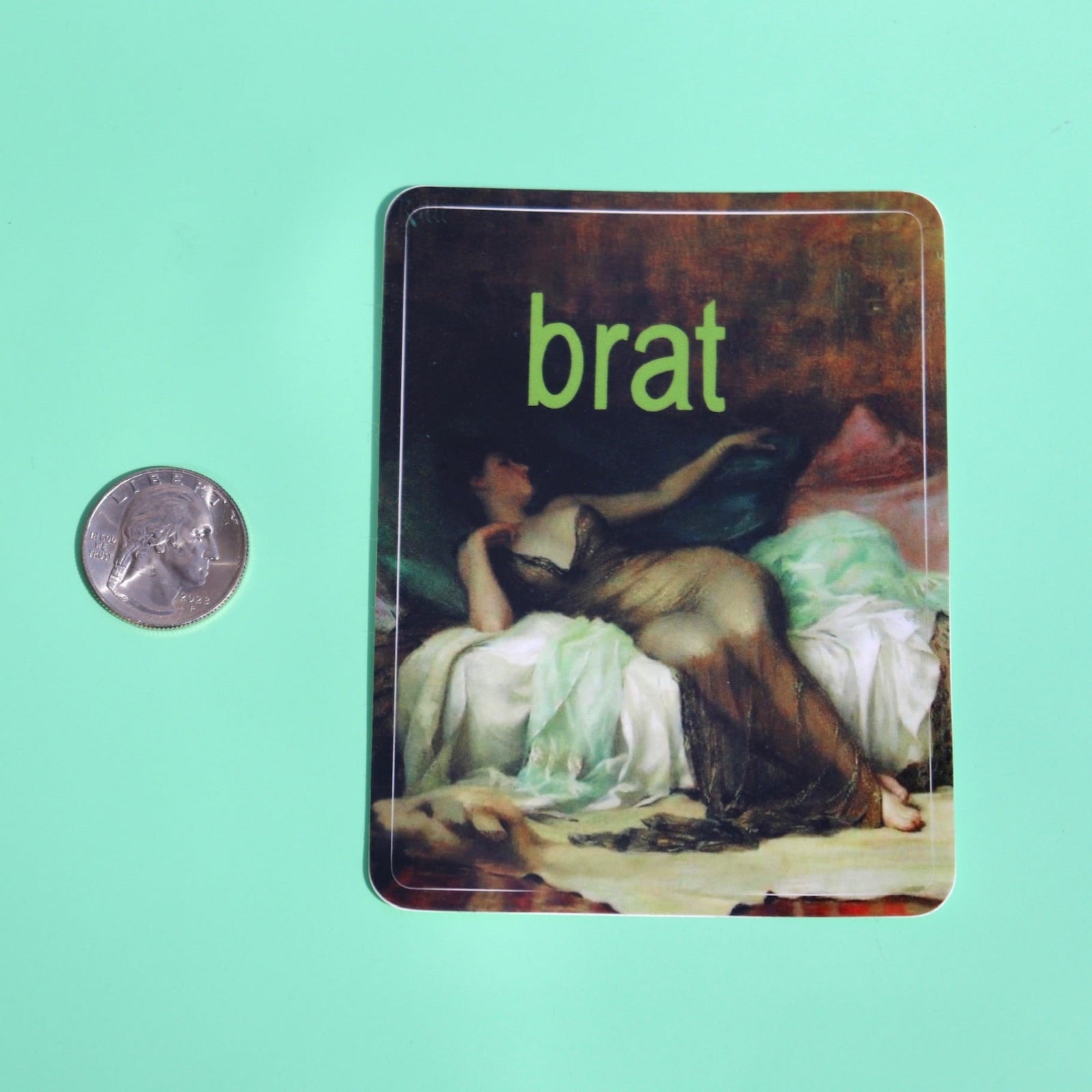 The Brat 3 Inch Vinyl Sticker next to a quarter on a green background.