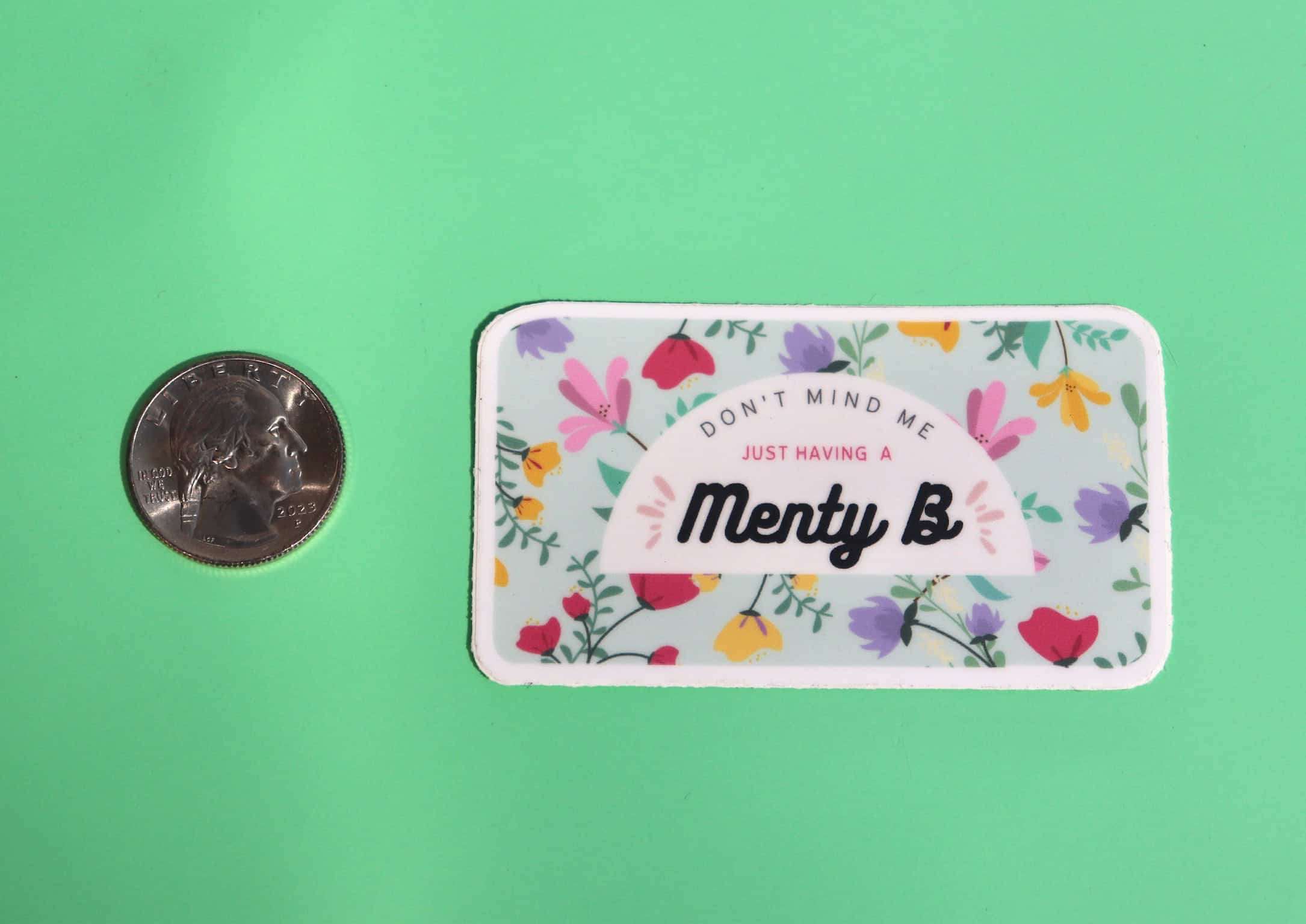 The Menty B Mental Health Anxiety Sticker next to a quarter on a green background.