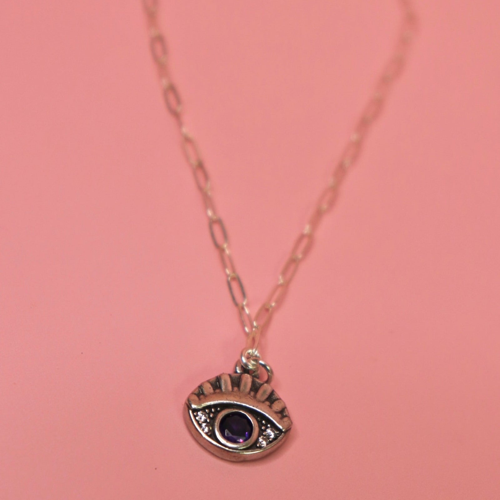 The Sterling Silver Purple Evil Eye Necklace with Paperclip Chain on a pink background.