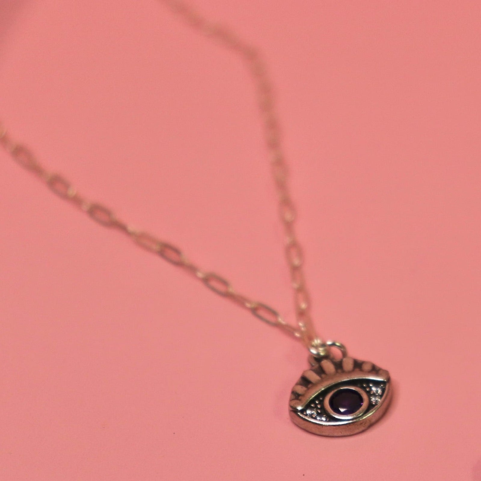 The Sterling Silver Purple Evil Eye Necklace with Paperclip Chain on a pink background.