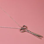 An up-close image of the Vintage Silver Scissors Charm Necklace on a pink background.