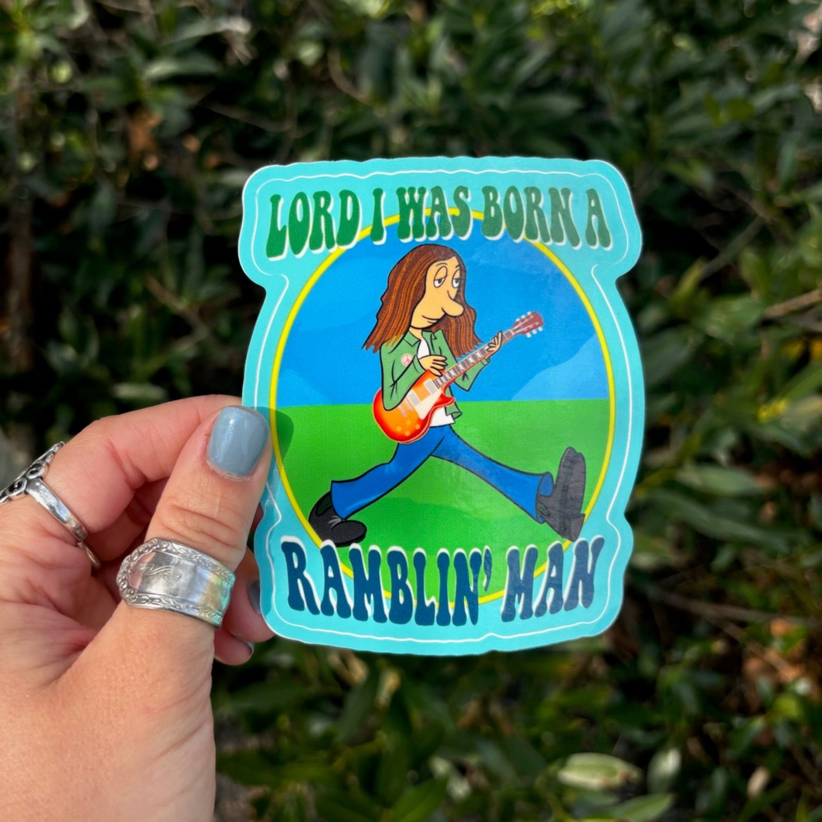 Someone holding the Ramblin' Man 3 Inch Sticker in front of greenery.