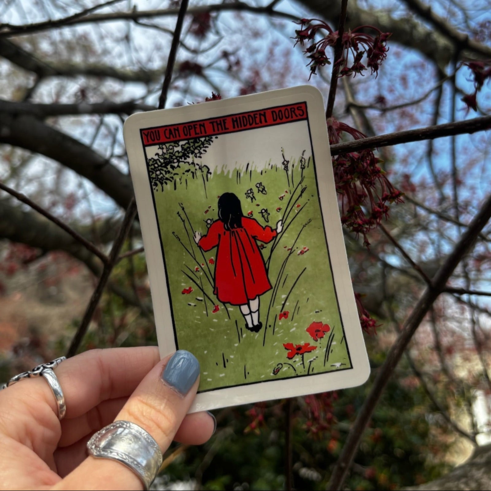 Someone holding the You Can Open The Hidden Doors 3 Inch Sticker in front of tree branches with flowers.