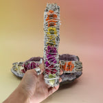 The 'Joy' Floral White Sage Smudge Stick being held in front of another 'Joy' Floral White Sage Smudge Stick on an amethyst piece all on a pink and yellow background.