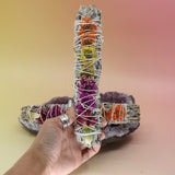 The 'Joy' Floral White Sage Smudge Stick being held in front of another 'Joy' Floral White Sage Smudge Stick on an amethyst piece all on a pink and yellow background.