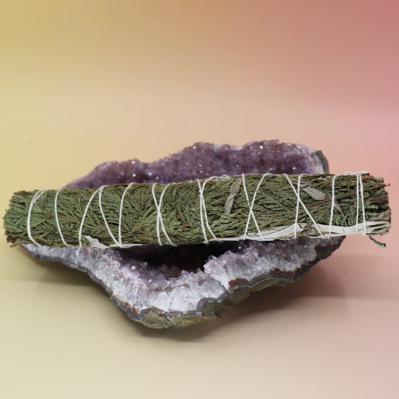 The White Sage & Cedar Smudge Stick 9 in. on an amethyst piece all on a pink and yellow background.