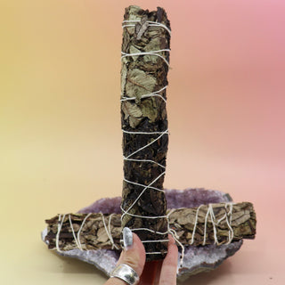 Someone holding the Black Sage Smudge Stick 9 in. in front of another Black Sage Smudge Stick 9 in. on an amethyst piece all on a pink and yellow background.