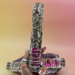 The Dahlia White Sage Smudge Stick 9 in. being held in front of another Dahlia White Sage Smudge Stick 9 in. on an amethyst piece all on a pink and yellow background.
