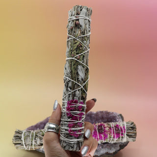The Dahlia White Sage Smudge Stick 9 in. being held in front of another Dahlia White Sage Smudge Stick 9 in. on an amethyst piece all on a pink and yellow background.
