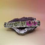 The Dahlia White Sage Smudge Stick 9 in. on an amethyst piece all on a pink and yellow background.