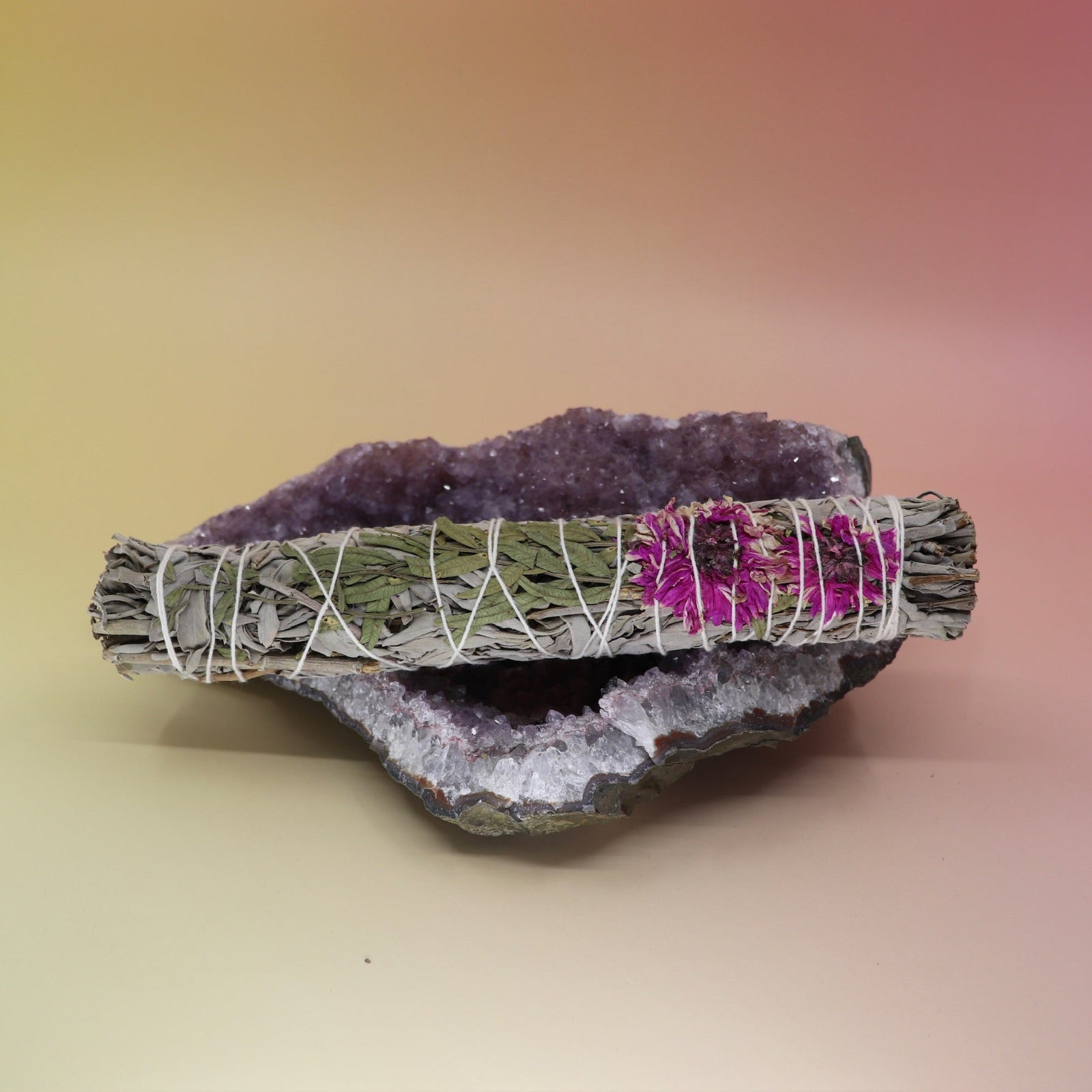 The Dahlia White Sage Smudge Stick 9 in. on an amethyst piece all on a pink and yellow background.