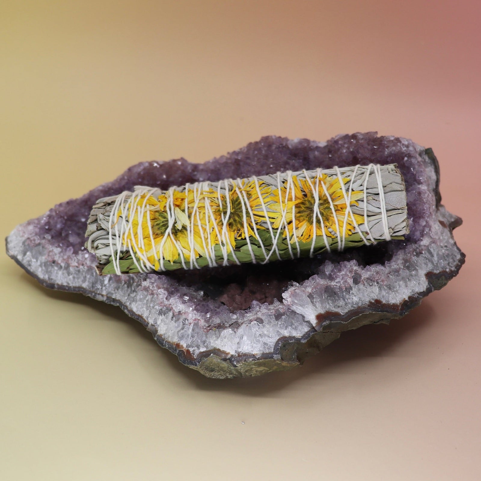 The Organic Sunflower White Sage  - 4 in. Smudge Stick on an amethyst piece all on a pink and yellow background.
