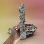 The White Sage & Juniper Smudge Stick 9 in. being held in front of another White Sage & Juniper Smudge Stick 9 in. on an amethyst piece all on a pink and yellow background.