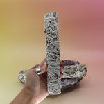 Someone holding the White Sage 9 in. Smudge Stick for Cleansing in front of an amethyst piece all on a pink and yellow background.