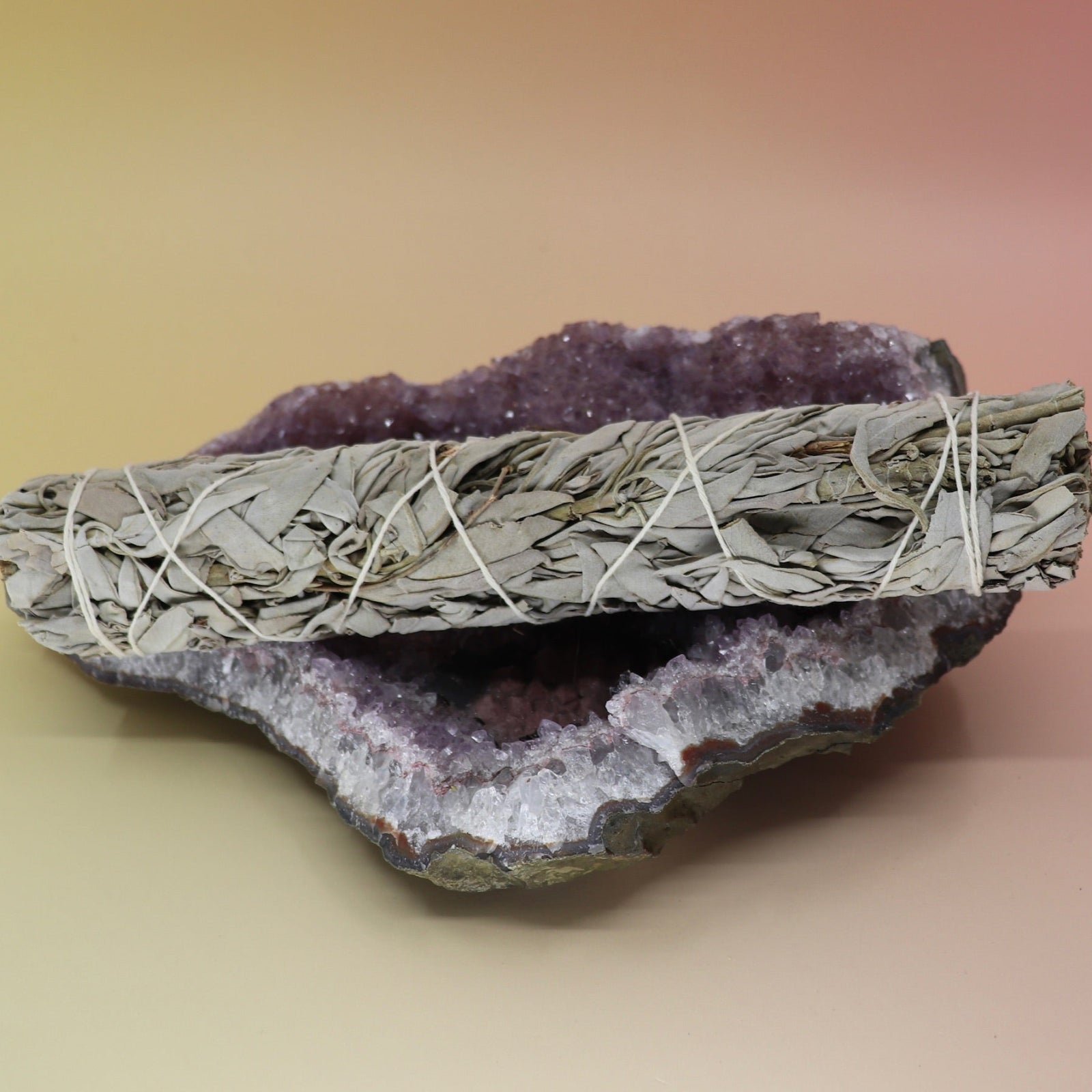 The White Sage & Juniper Smudge Stick 9 in. on an amethyst piece all on a pink and yellow background.