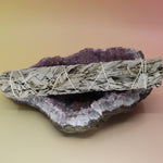 The White Sage 9 in. Smudge Stick for Cleansing on an amethyst piece all on a pink and yellow background.