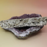 The White Sage 9 in. Smudge Stick for Cleansing on an amethyst piece all on a pink and yellow background.