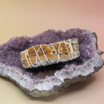 The Organic White Sage with Cinnamon & Orange - 4 in. Smudge Stick Bundle on an amethyst piece all on a pink and yellow background.