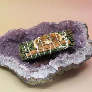 The Organic Cedar, Orange & Cinnamon - 4 in. Smudge Stick Bundle on an amethyst piece all on a pink and yellow background.