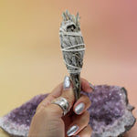 The California White Sage Torch 4 in. being held in front of an amethyst piece on a pink and yellow background.