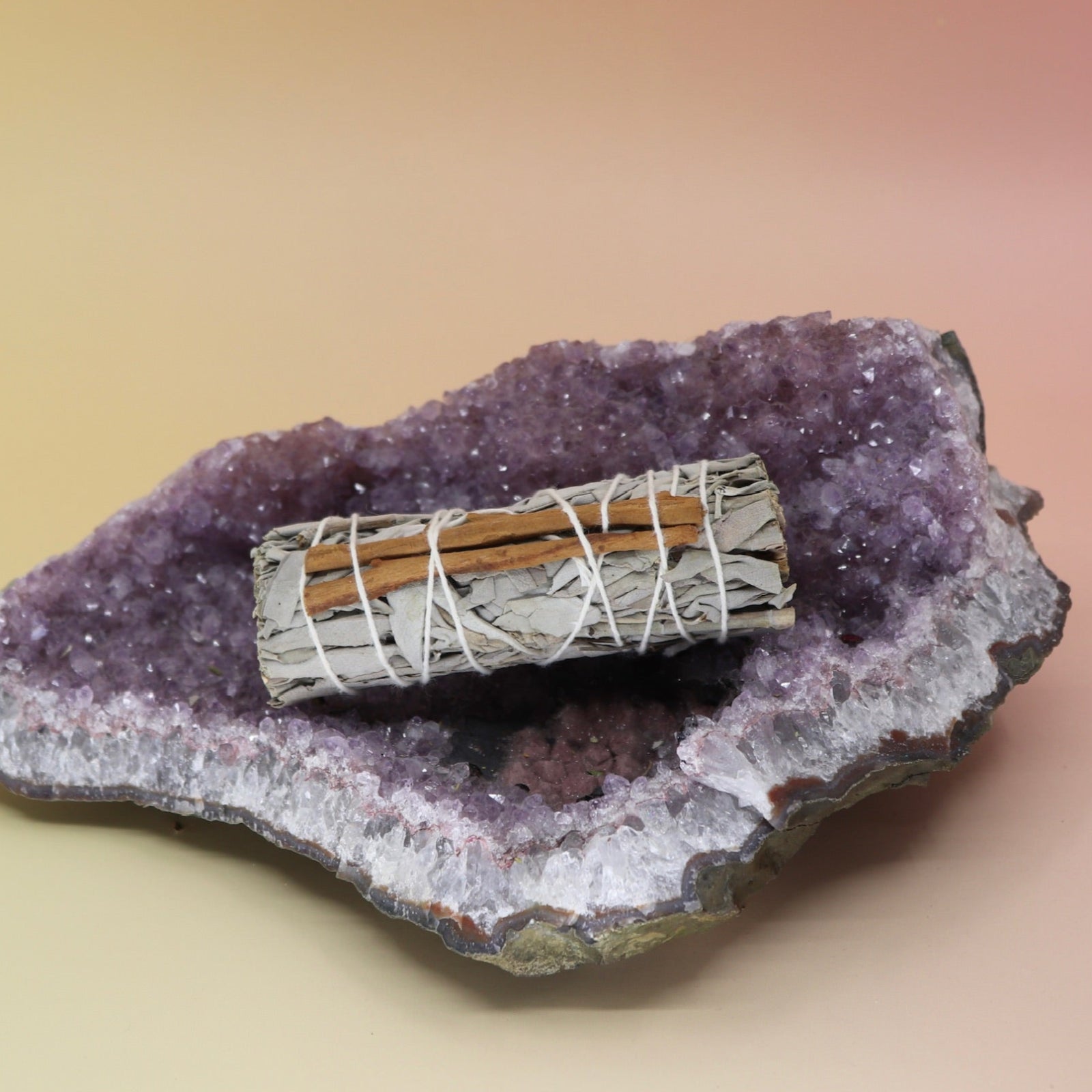 The Organic White Sage & Cinnamon - 4 in. Smudge Stick Bundle - Love, Luck and Money Drawing Smudge on an amethyst piece all on a pink and yellow background.
