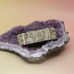 The Organic Blue Sage - 4 in. Smudge Stick on an amethyst piece all on a pink and yellow background.
