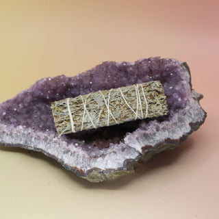 The Organic Blue Sage - 4 in. Smudge Stick on an amethyst piece all on a pink and yellow background.