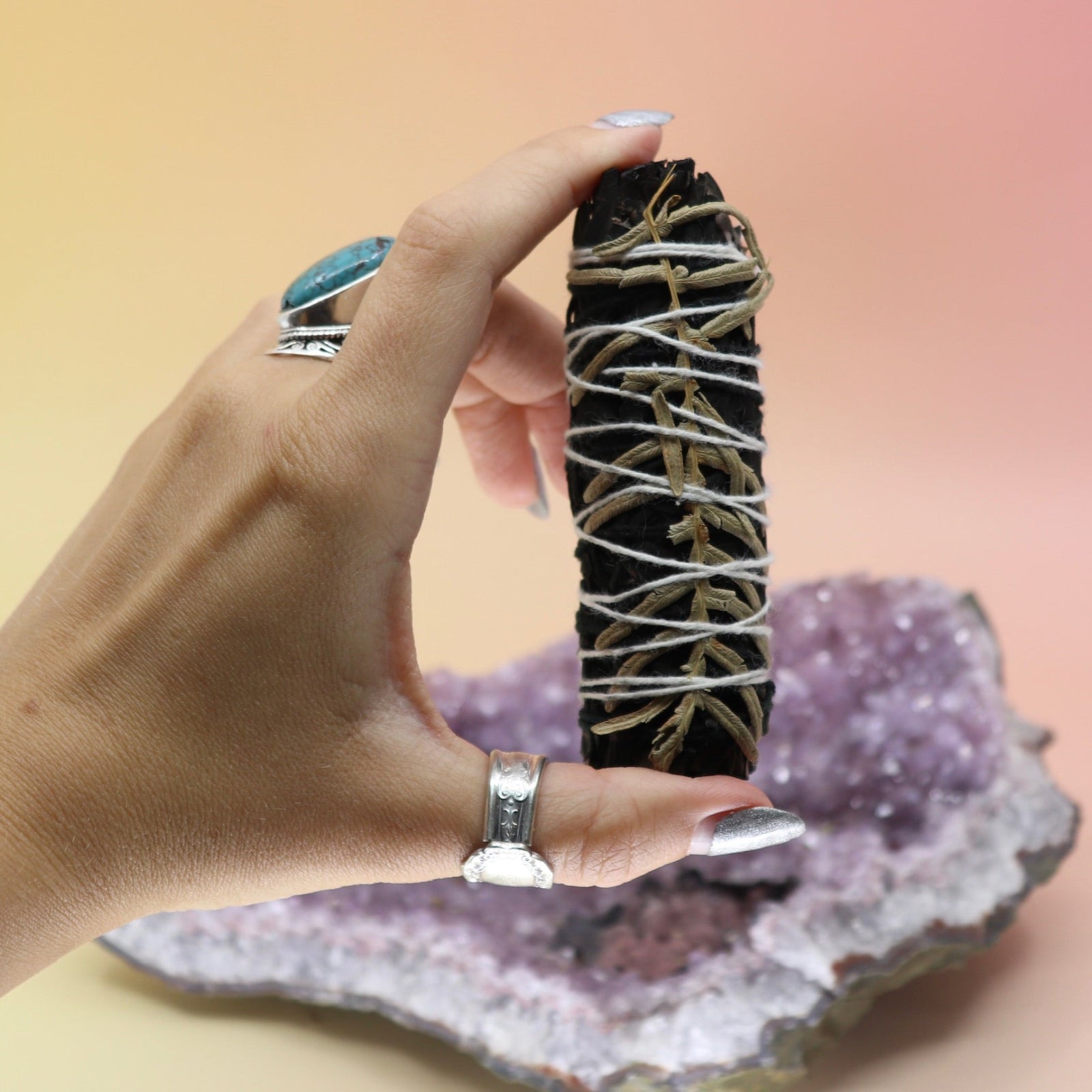 Someone holding the 'Terra' Organic Black Colored White Sage with Cream Leaves - 4 in. Smudge Stick in front of an amethyst piece all on a pink and yellow background.