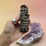Someone holding the 'Terra' Organic Black Colored White Sage with Cream Leaves - 4 in. Smudge Stick in front of an amethyst piece all on a pink and yellow background.
