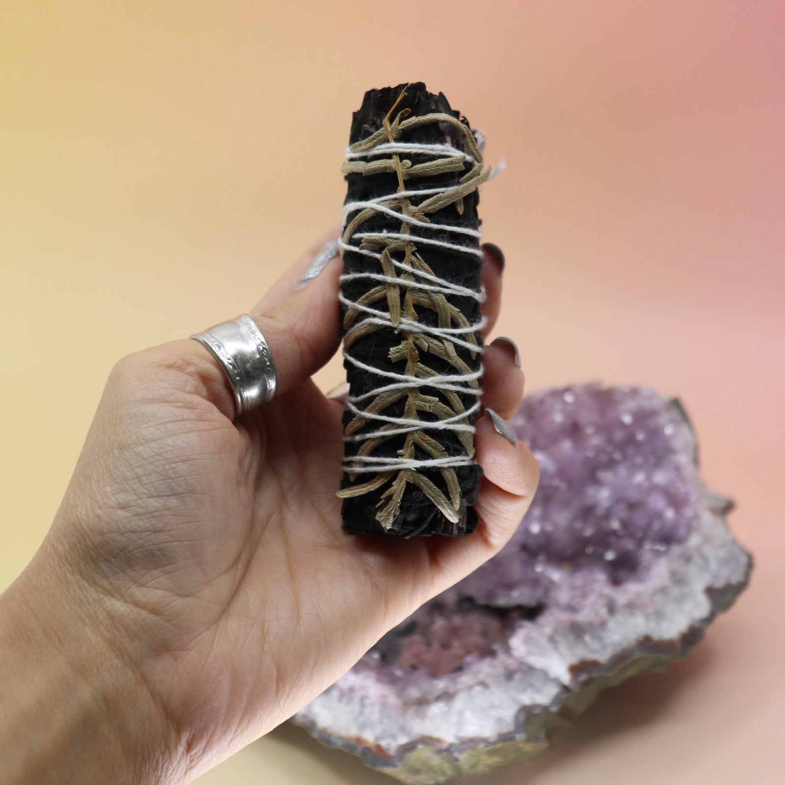 Someone holding the 'Terra' Organic Black Colored White Sage with Cream Leaves - 4 in. Smudge Stick in front of an amethyst piece all on a pink and yellow background.