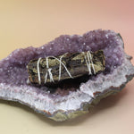The Organic Black Sage - 4 in. Smudge Stick on an amethyst piece all on a pink and yellow background.