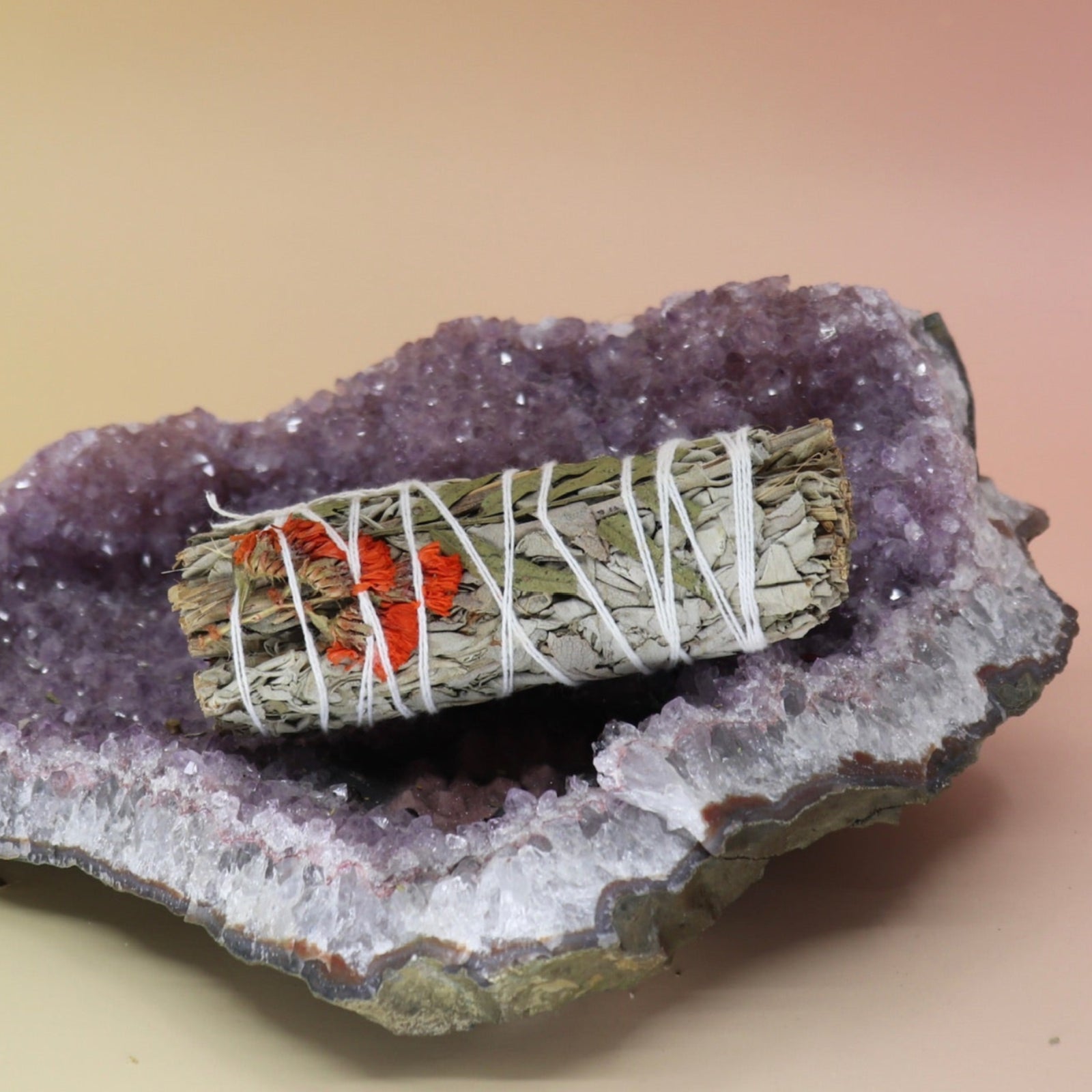 The 'Positive Vibes' Floral Sage - 4 in. Smudge Stick on an amethyst piece all on a pink and yellow background.