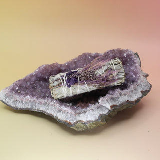 The 'Purple Bouquet' Organic Floral White Sage with Purple Flowers - 4 in. Sage Smudge Stick on an amethyst piece on a pink and yellow background.
