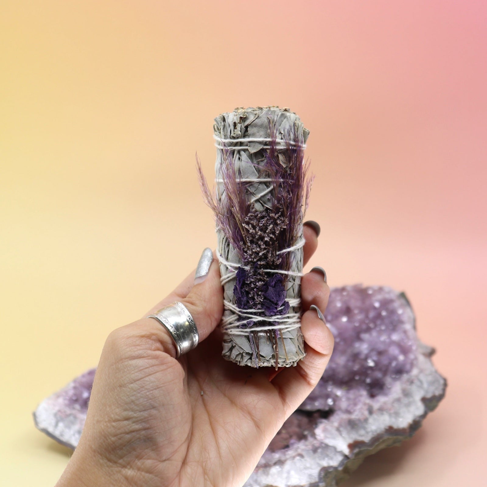 Someone holding the 'Purple Bouquet' Organic Floral White Sage with Purple Flowers - 4 in. Sage Smudge Stick in front of an amethyst piece on a pink and yellow background.
