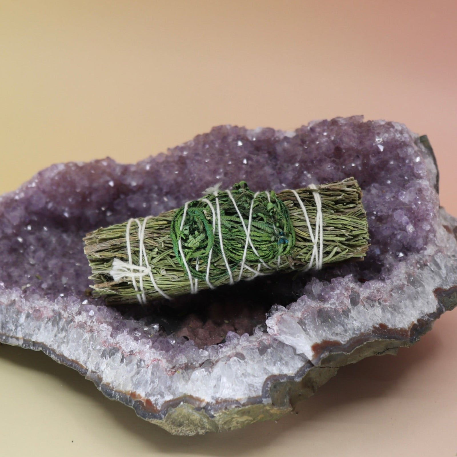 The 'Jericho Dreams' Organic Floral Rosemary Sage with Jericho Flower - 4 in. Smudge Stick on an amethyst piece all on a pink and yellow background.