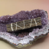 The Organic Copal Sage 4 in. Smudge Stick on an amethyst piece all on a pink and yellow background.