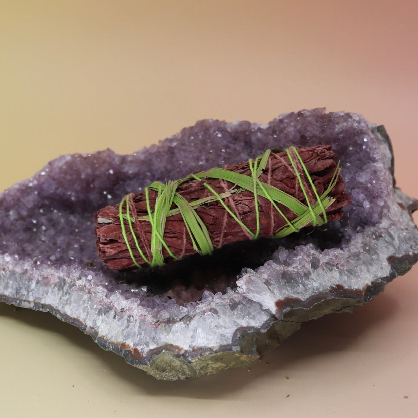 The Secret Garden Floral Dragons Blood Sage - 4 in. Smudge Stick on an amethyst piece all on a pink and yellow background.