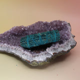 The Nag Champa Scented Sage Smudge Stick 4 in. on an amethyst piece on a pink and yellow background.