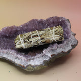 The Organic Yerba Santa Sage - 4 in. Smudge Stick on an amethyst piece all on a pink and yellow background.
