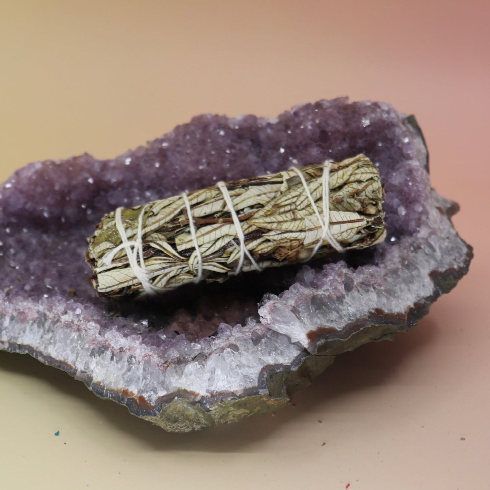 The Organic Yerba Santa Sage - 4 in. Smudge Stick on an amethyst piece all on a pink and yellow background.