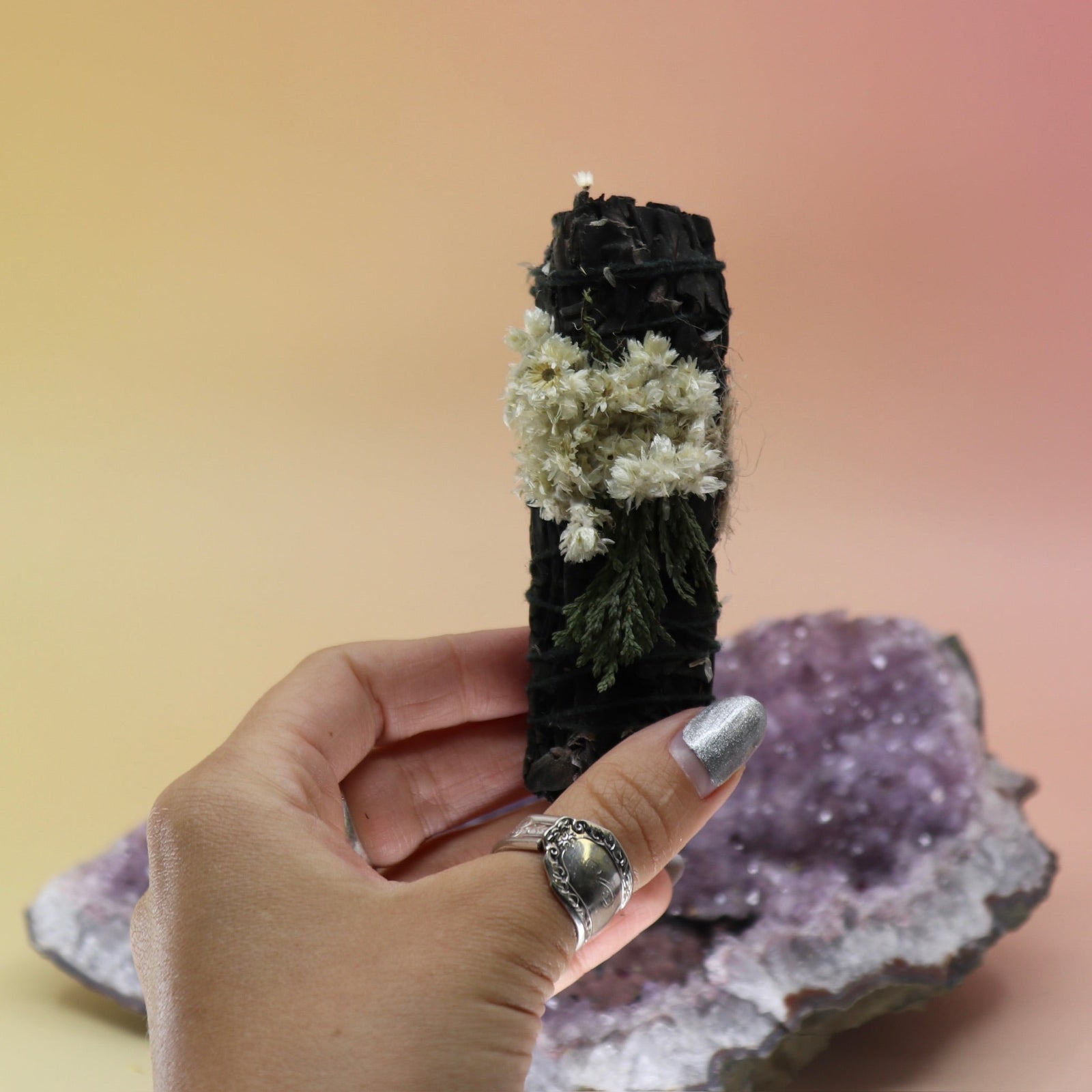The 'Midnight Bloom' Floral White Sage Smudge Stick 4 in. being held in front of an amethyst piece on a pink and yellow background.