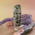 Someone holding the Organic White Sage & Yerba Santa - 4 in. Smudge Stick in front of an amethyst piece on a pink and yellow background.