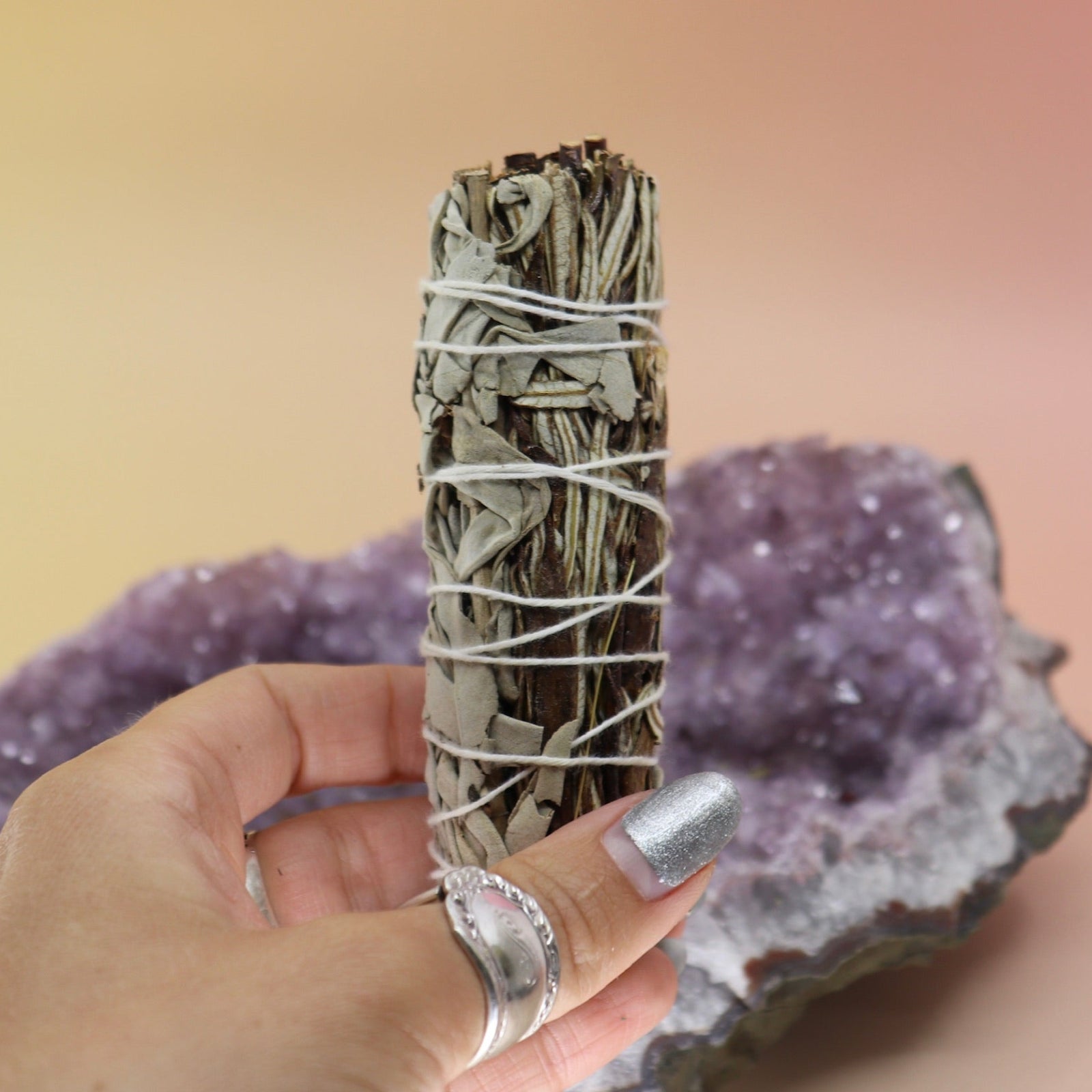 Someone holding the Organic White Sage & Yerba Santa - 4 in. Smudge Stick in front of an amethyst piece on a pink and yellow background.